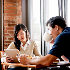 Two people dining and looking at electronic devices, with windows in the background.