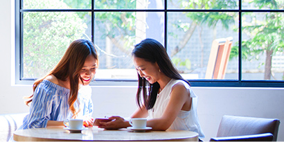 Two women smiling over a phone in a sunny restaurant with many windows.
