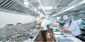 A long, narrow commercial kitchen with stainless steel finishes has several chefs in white busily preparing food.
