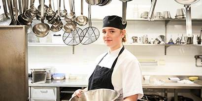 A smiling kitchen worker stands among hanging kitchen tools, including ladles and strainers.
