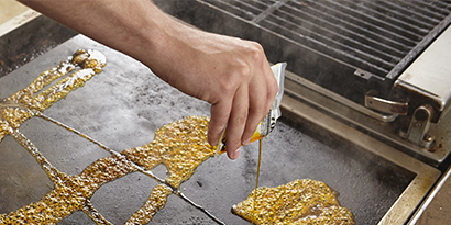 A commercial kitchen worker pours Scotch-Brite™ Quick Clean Griddle Liquid onto a flat-top griddle.
