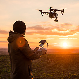 A man outside using a two-handed remote to control a drone with a camera in the sky.