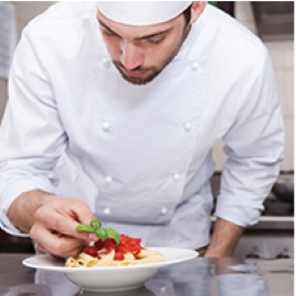 A chef carefully adds an accent to a bowl of pasta in a commercial kitchen.