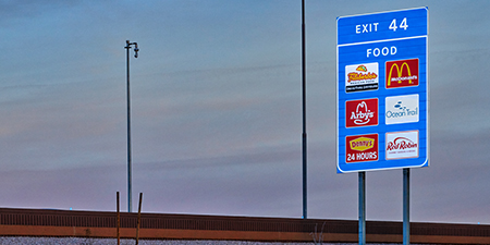 A blue exit sign listing restaurants in front of a dusk sky.
