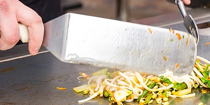 Closeup of two hands cooking vegetables on a flat-top grill.
