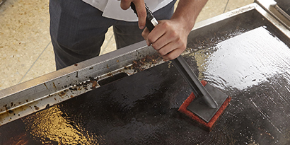 A worker cleans a flat-top griddle with Scotch-Brite™ Quick Clean Heavy Duty Griddle Pad and Multi-Purpose Pad Holder.

